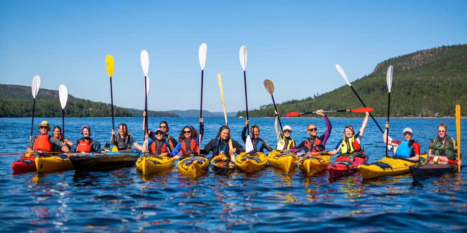14 people in kayaks, with their padel above their heads, smiling towards the photographer and the sun.