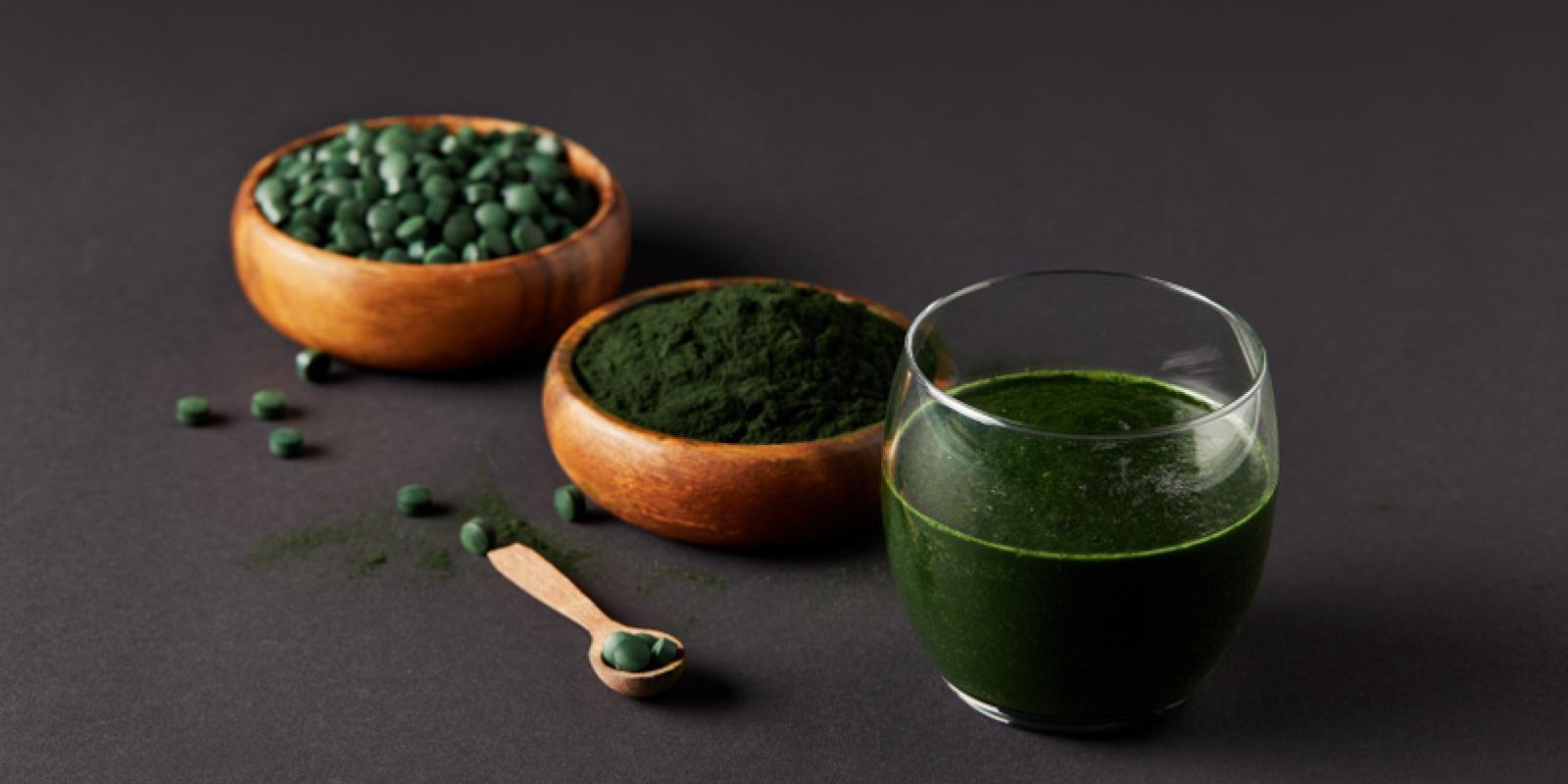 Trekopp med fersk smoothie av spirulina, bolle med spirulinapulver og spirulinapiller på et grått bord.