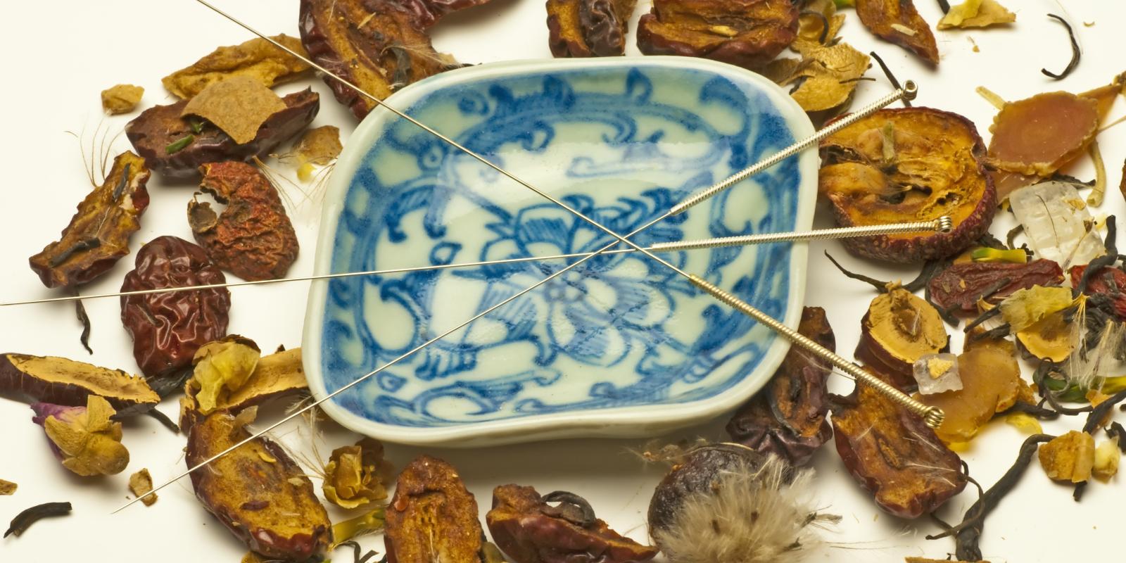 Bilde av akupunkturnåler som ligger oppå en skål med asiatiske skrifttegn og av tørkede urter ligger rundt skålen