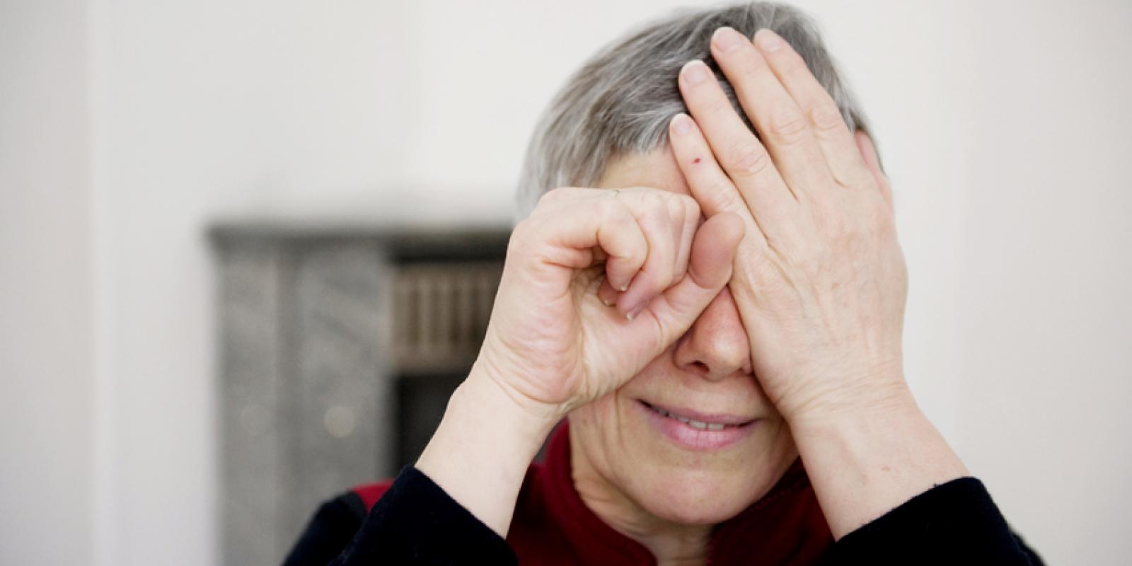 Kvinne med grått, kort hår, holder seg for et øye og lager kikkert med den andre hånden foran det andre øyet.